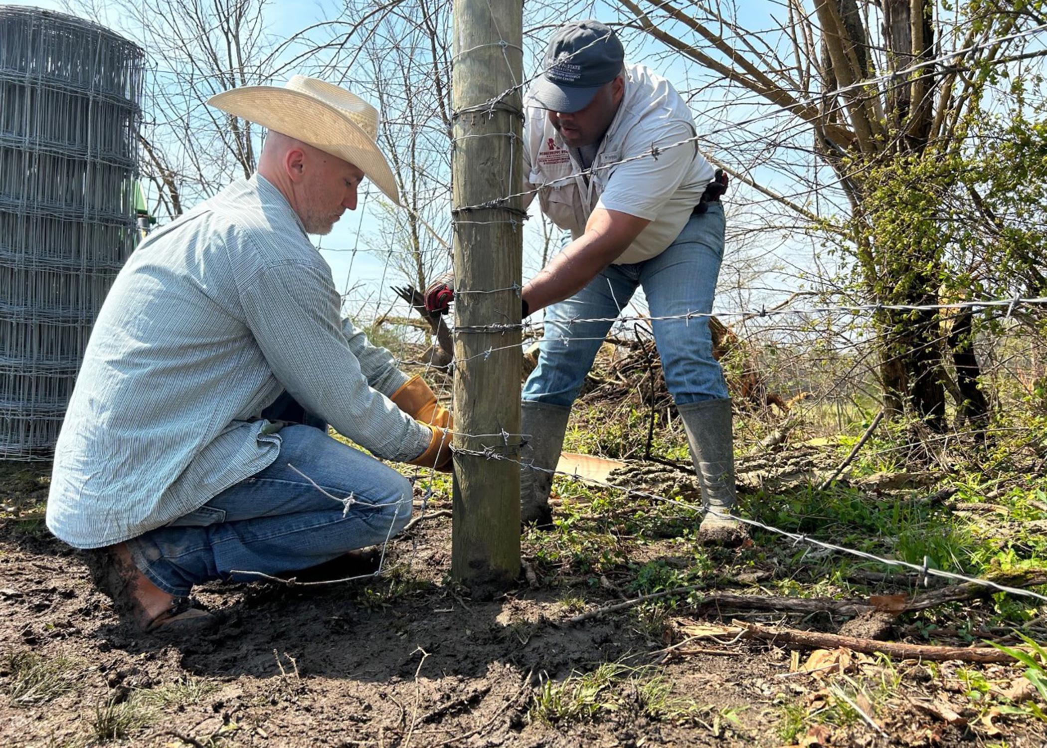 Two men tie barbed wire around a fence post.