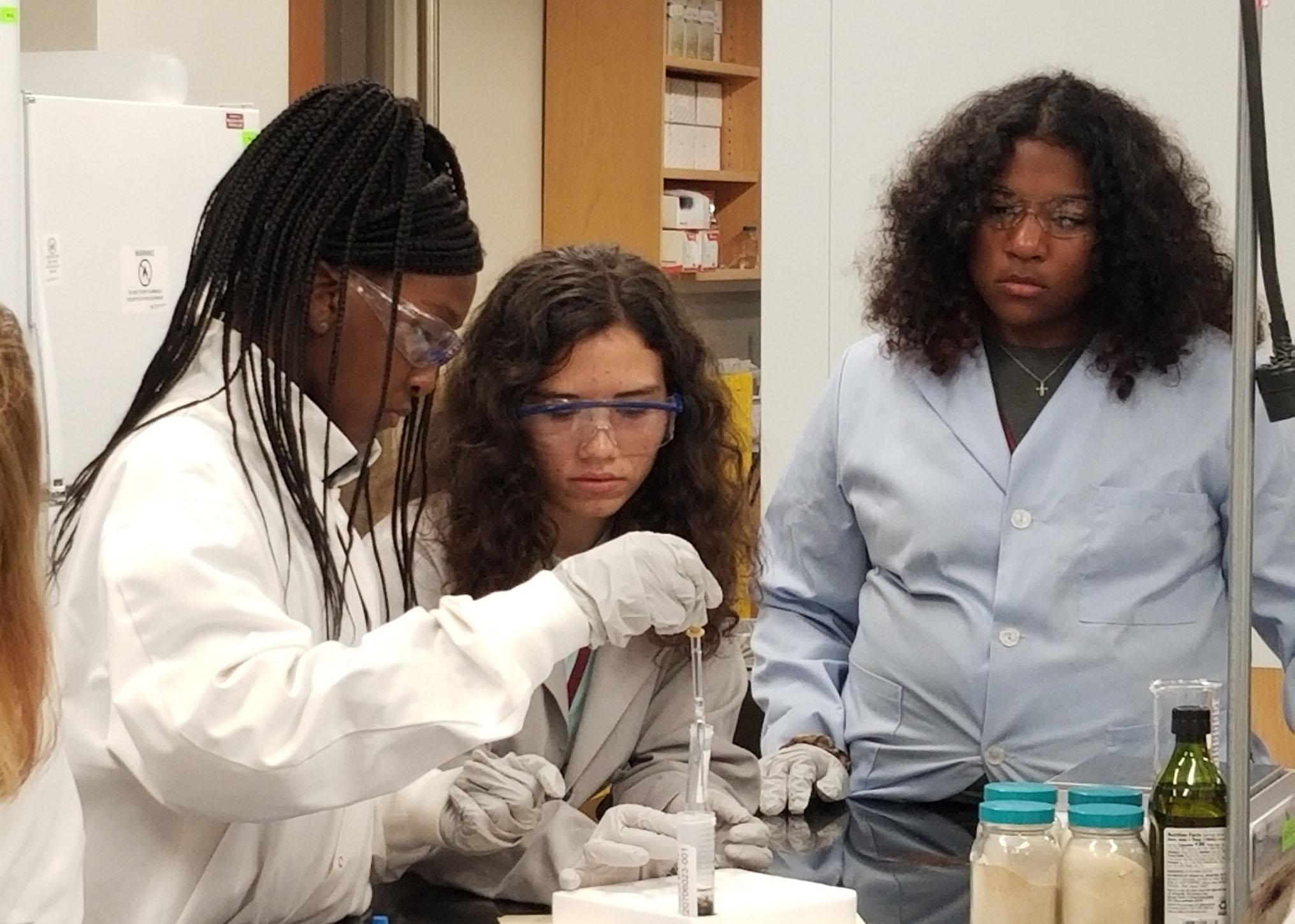 Three girls in lab coats work with scientific equipment.