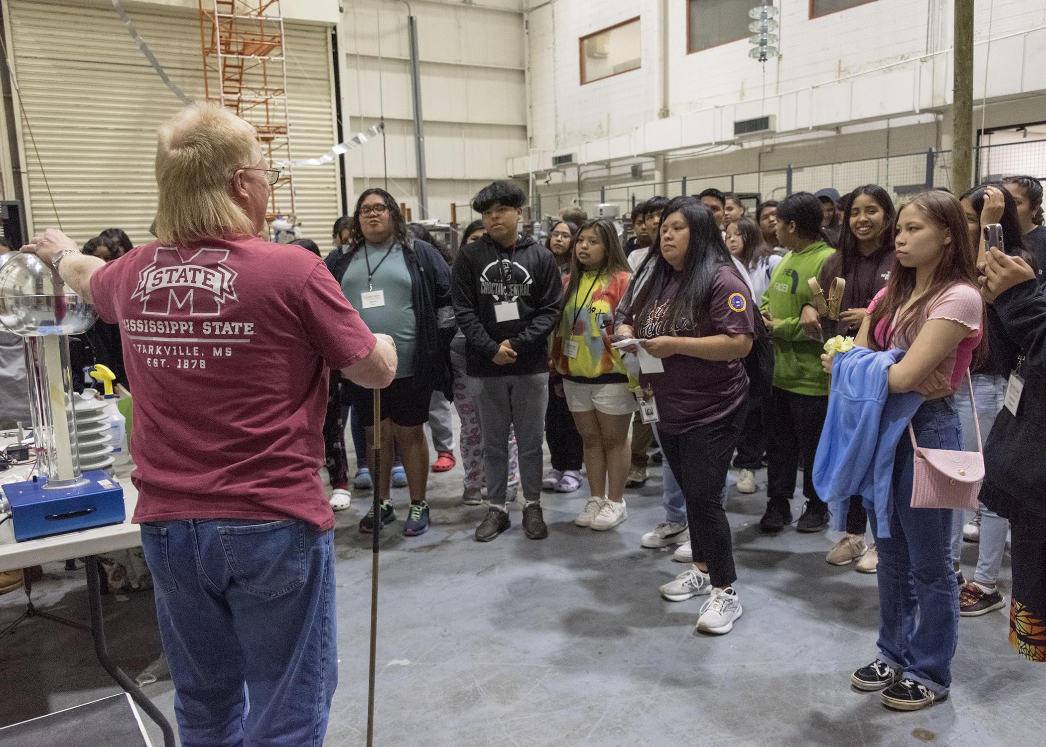 A man shows a Van de Graaff generator to students on a class trip.	