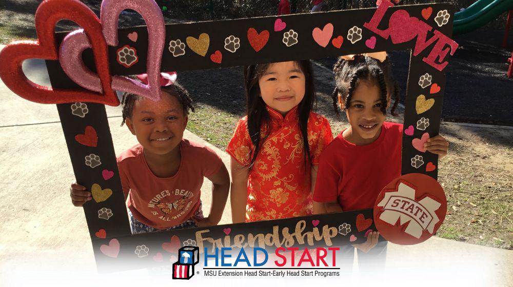 Young students with smiling faces holding up a Valentine's Day sign.