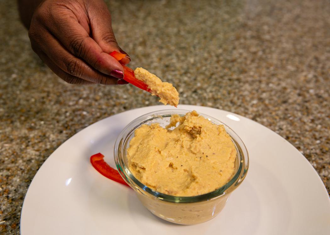 A hand is shown dipping a bell pepper strip into a bowl of garlic chickpea dip.