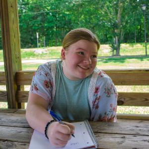 A girl smiling and looking up from her notepad.