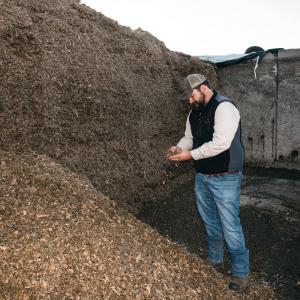 A man holding a handful of mulch standing beside a hill of mulch.