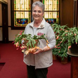 An older woman holding a potted Christmas cactus with peach blooms in front and pink blooms in back.