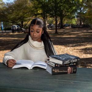 A Black woman wearing a white turtleneck sweater looks at large, thick books.