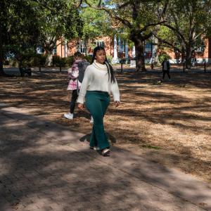 A Black woman wearing a white turtleneck sweater walks along a path outside.