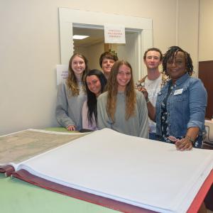 Teens standing beside a woman looking at a large book.