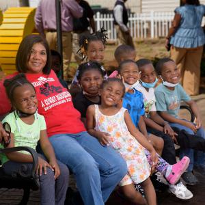 A woman, wearing a red T-shirt listing Rosemary’s Daycare on it, smiles while sitting on a bench with many small children sitting beside her, also smiling.