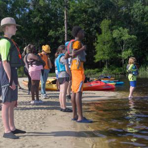 A group of children standing on sand and wearing lifejackets, looking out across water.