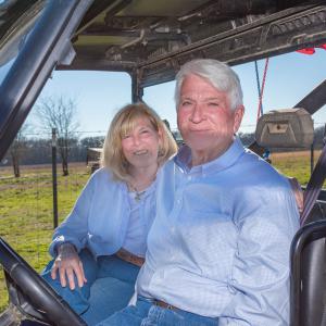 A woman and man seated in a side-by-side with a cattle field stretching behind them.