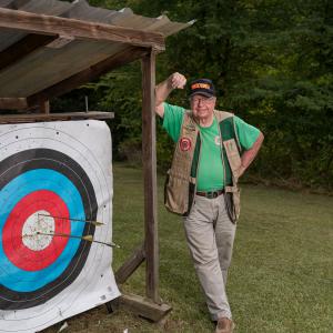 A smiling man wearing a vest and green shirt, standing beside an archery target with three arrows near the bullseye.