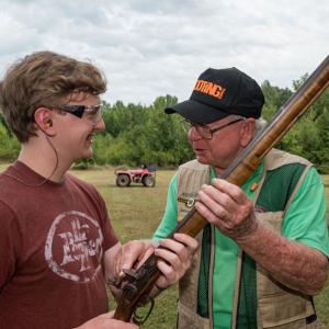 A man wearing a vest and green shirt, talking to a young man smiling and holding a shotgun.