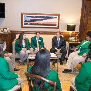 Seven teens in green blazers sit on chairs and couches to listen to a man with glasses and wearing a black suit and blue tie.
