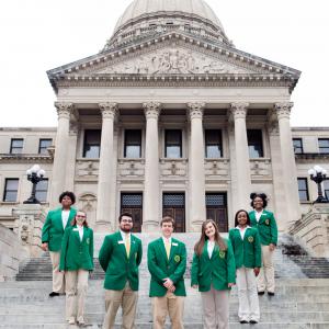 Seven teenagers wearing green blazers and tan slacks form a U standing on steps in front of six gray columns.