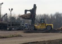 A yellow piece of heavy machinery lifts a load of cut trees off the back of a log truck in a sawmill yard.