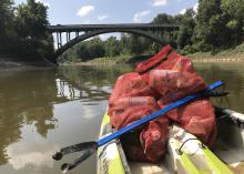 The front of a kayak has two red meshed bags containing litter and a blue trash-grabbing tool with the words “Pearl Riverkeeper” printed on it. The boat is on a small river and approaching a highway bridge.