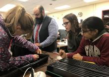 Two middle-school-aged girls and a middle-aged male teacher look on at another girl poking a hole in the side of a black tray for a science project.