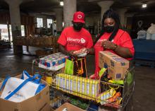 Two people in red shirts stand behind a large basket of grocery items.