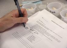 A hand with a pen rests on a paper next to numbered sample cups.