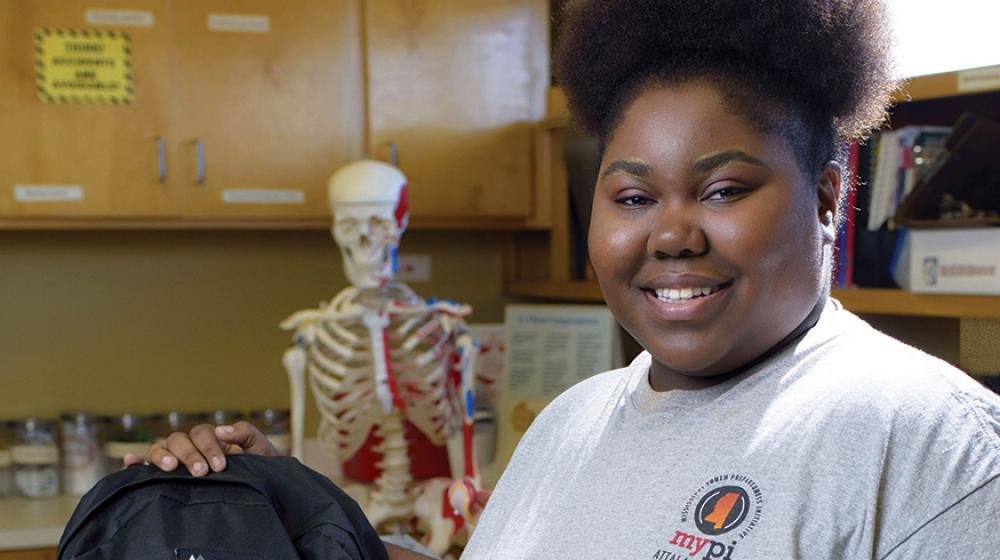 A smiling teenage girl standing next to a black backpack.