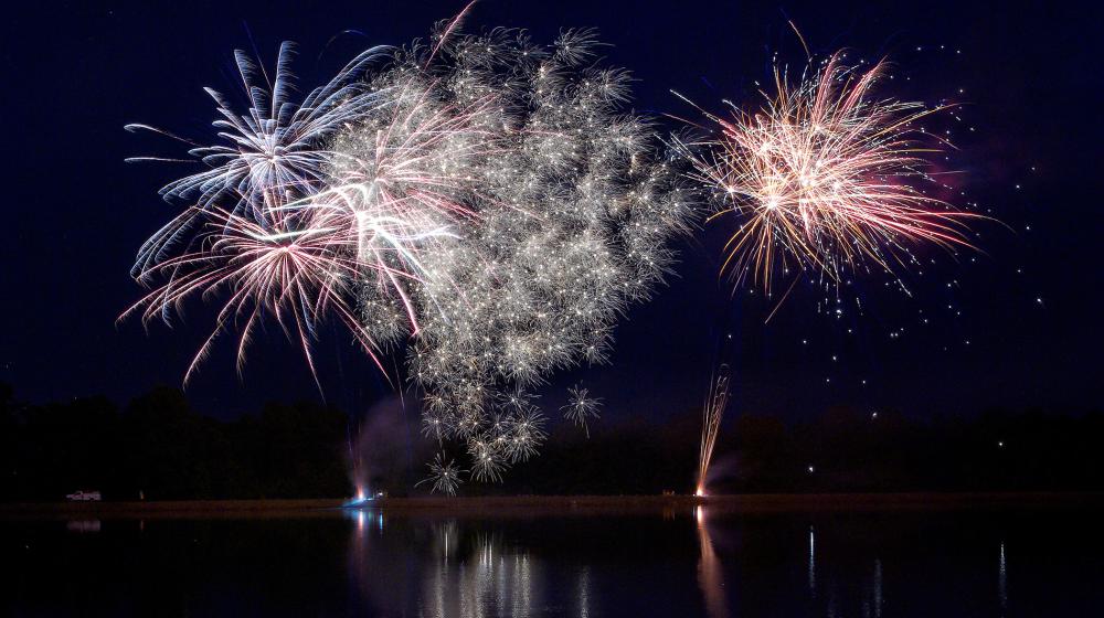 Fireworks explode against a dark sky and above a large body of water. 