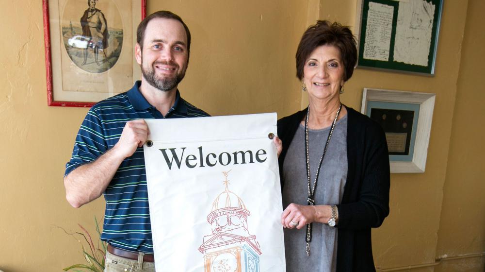 A man in a blue shirt and a woman in a grey shirt with a black cardigan stand holding a banner that says “Welcome…the Square Kosciusko.”