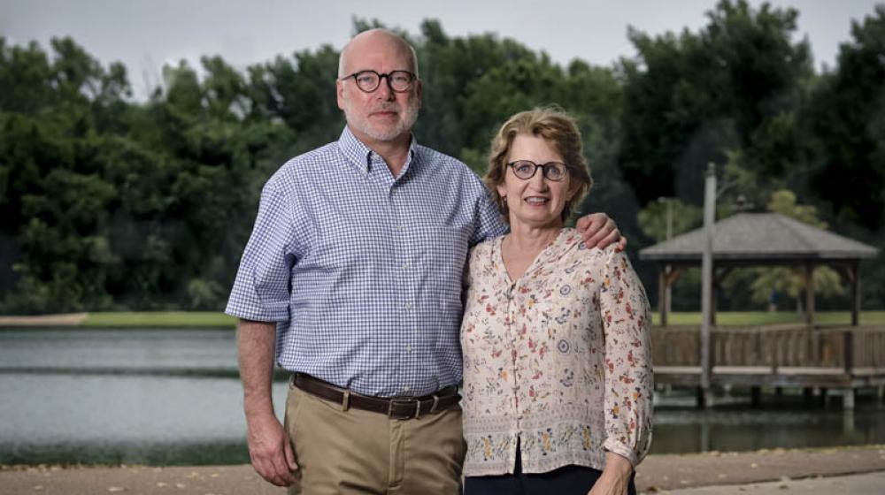 A man with his arm around a smiling woman standing on a sidewalk in front of a lake.