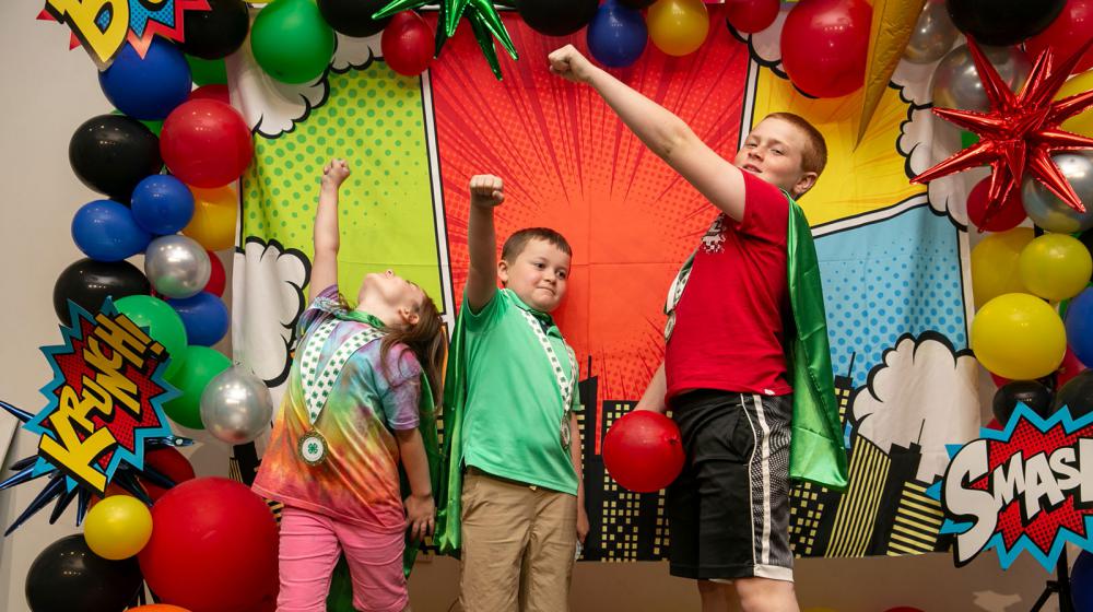 Three children, wearing capes and standing with fists raised.