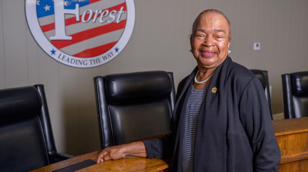 A woman stands smiling in front of the seal for Forest, MS, that lists “Leading the Way” across the bottom.