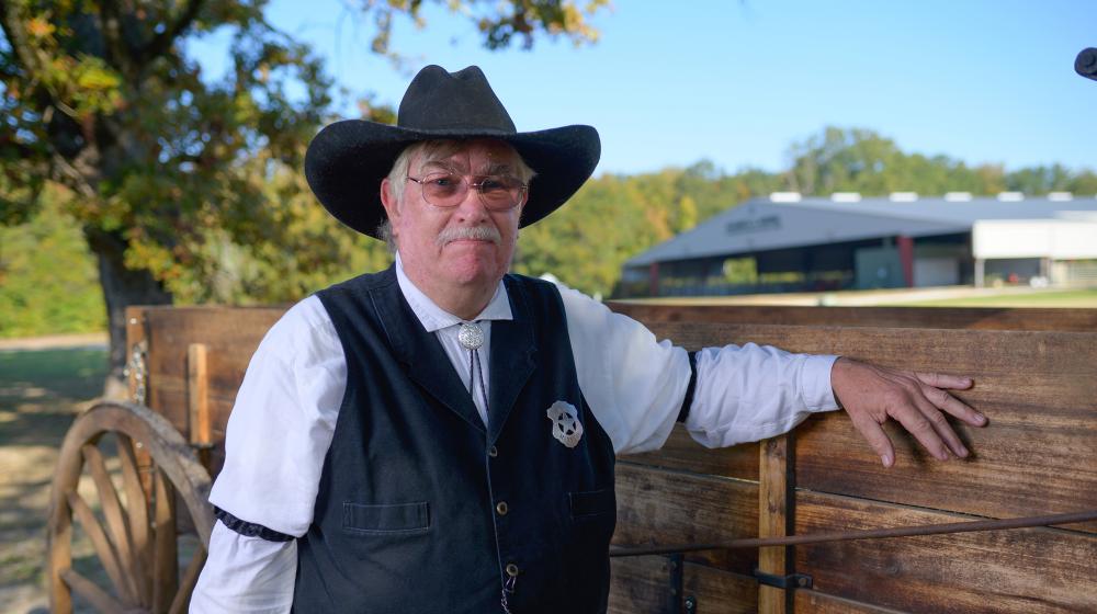 A person with a gray mustache and wearing a black cowboy hat, white polo, and black vest with a sheriff’s star on his chest.