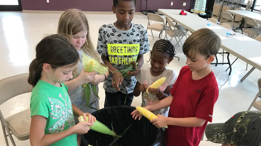 Children stand around a large garbage can while they shuck the husks and silks from ears of corn. 