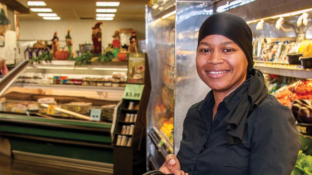 A woman in a grocery store holding a blue basket filled with produce like leafy green lettuce.