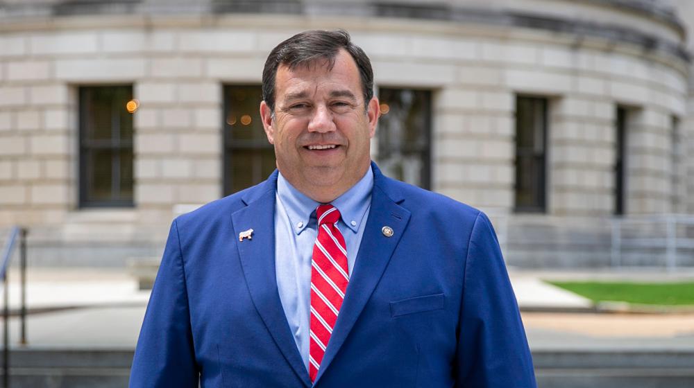 A man, smiling, standing outside and wearing a blue suit with a white-striped red tie.