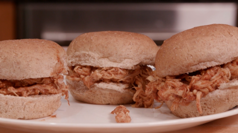 Three small sandwiches made with whole wheat slider buns, shredded chicken, and barbecue sauce on a white plate. 