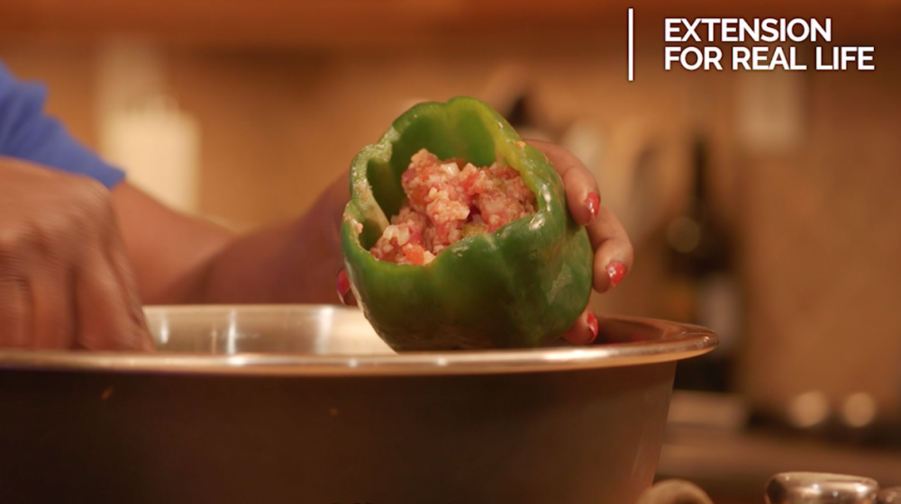 Hands spoon a ground meat and riced cauliflower mixture into a raw green bell pepper held over a stainless steel bowl.