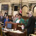 A young woman wearing a dark green blazer stands behind a podium while several people stand looking at her. 