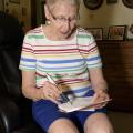 A gray-haired woman in blue shorts and a multi-colored striped shirt holds a pen over a pocket calendar to check appointments.