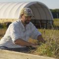 A man in a baseball cap reaches into a patch of grass, while a tractor and a white, high-tunnel structure stand behind him.