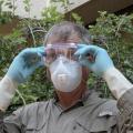 A man in a brown shirt, safety mask and rubber gloves adjusts a pair of goggles over his eyes.