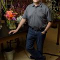 A man rests his hand on a table displaying floral arrangements.