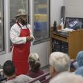 A man in a red apron speaks to a class in a lab.