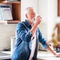 An older man drinks water in the kitchen.