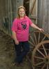 Dressed in a pink T-shirt and blue jeans, broiler grower Teresa Dyess stands next to two wagon wheels in front of a barn on her family farm.