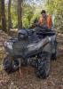 A hunter in camouflage and an orange vest places his rifle into storage on the back of an ATV in the woods.