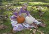 Two young girls sit on a colorful quilt among leaves in the grass as they play with a white and an orange pumpkin.