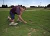 A hat rests on the ground next to a man kneeling down to examine grass.