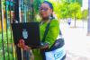 Woman in a green dress with a bag around her shoulder stands in front of a gate holding a laptop computer.