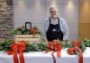 A man stands in a studio with a completed velvet bow, garland, door swag, and table centerpiece.