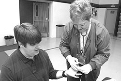 Woman wearing a stethoscope helping a man who is sitting down beside her.