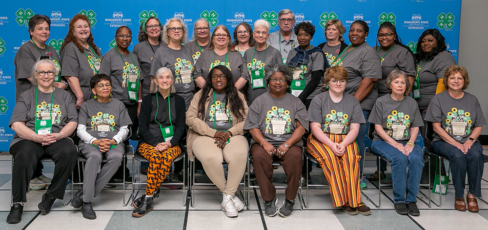 A group of people wearing a gray shirt with flowers standing in front of a blue wall with MSU Extension and 4-H logos in the background